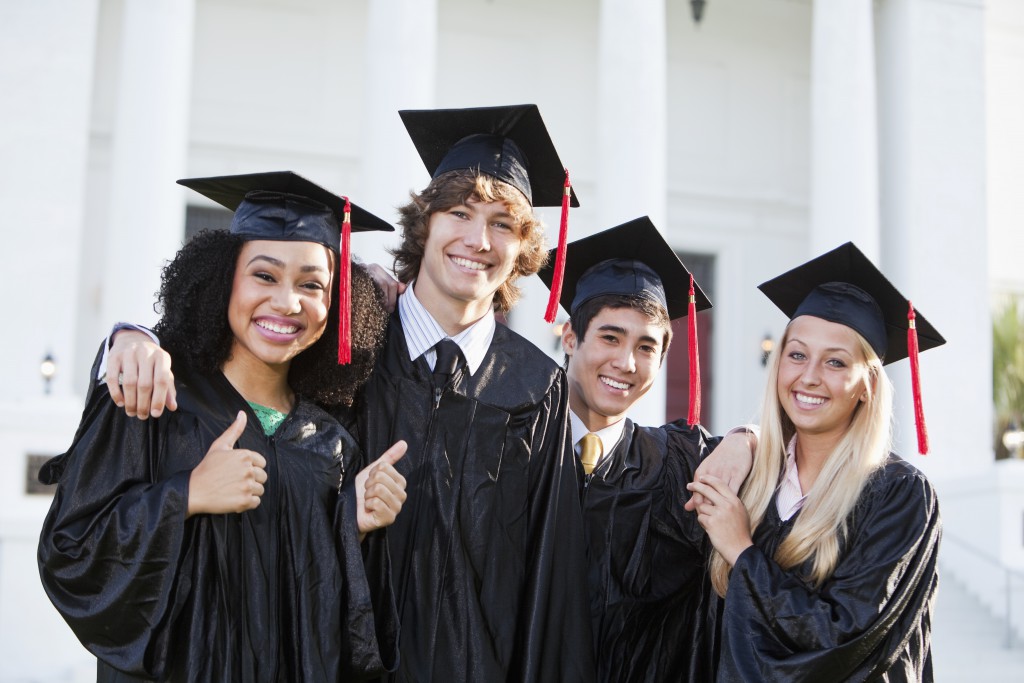 Multi-ethnic friends graduating together, in cap and gown in front of school building.